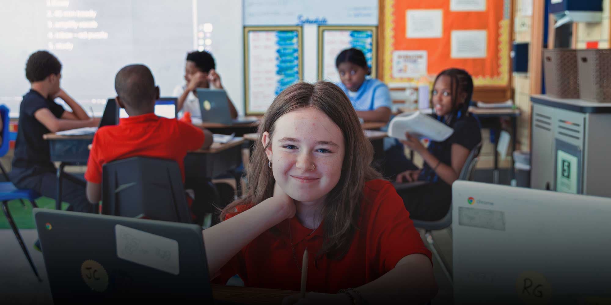 Smiling student at a desk with a laptop while classmates work in the background.