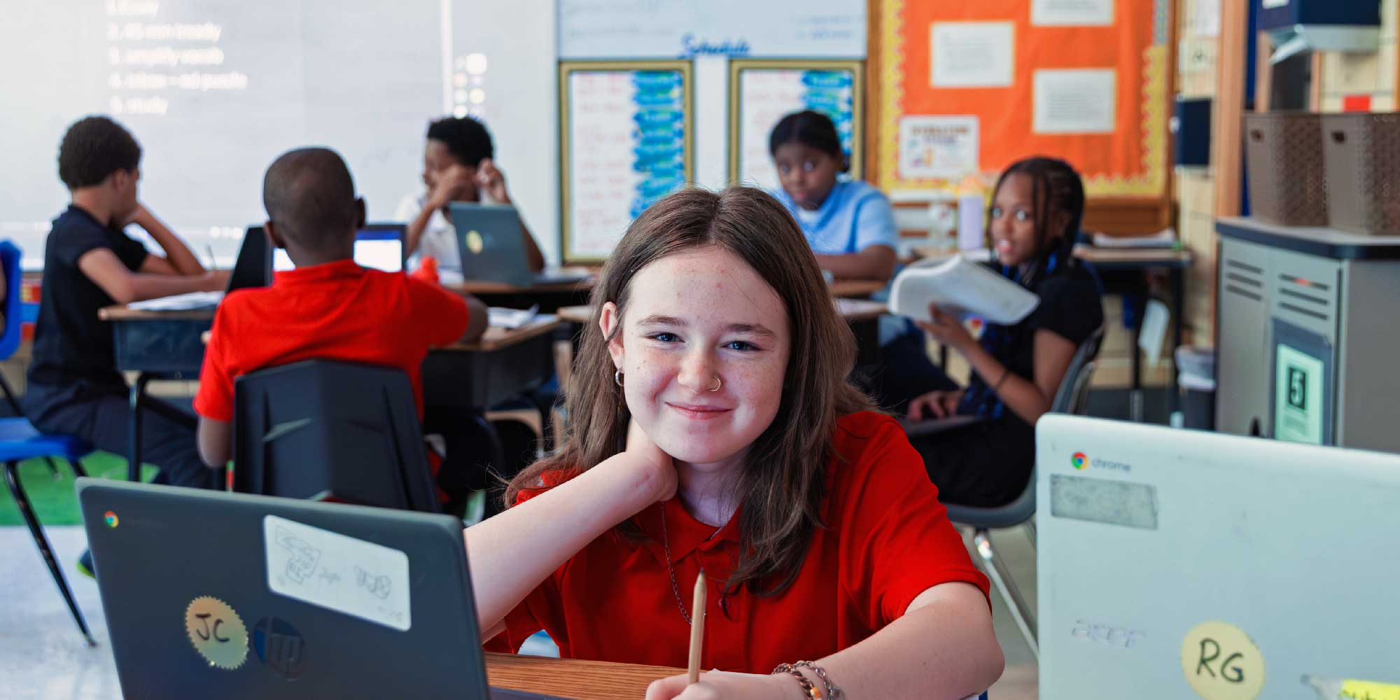 Smiling student at a desk with a laptop and pencil in a classroom.
